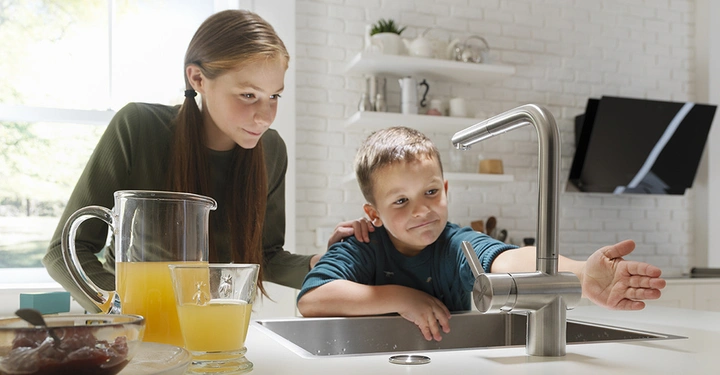Junge steht an einer Küchenspüle und hält seine Hand unter einen Wasserhahn Mädchen steht neben ihm Karaffe und Glas mit Orangensaft auf der Theke Weiße Küche mit Regalen im Hintergrund