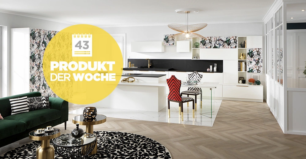 Wohnzimmer mit grüner Couch und schwarzen Kissen steht neben zwei goldenen runden Tischen mit Dekoration vor einem schwarz-weißen Teppich. Im Hintergrund moderne weiße Küche mit floralem Muster. Text: 43 PRODUKT DER WOCHE