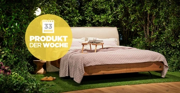 Ein Bett mit Holzrahmen und gemustertem Überwurf steht auf Rasen in einem von Pflanzen umgebenen Raum. Ein hölzernes Tablett mit Tassen steht auf dem Bett. Text: Kalender 33 PRODUKT DER WOCHE.