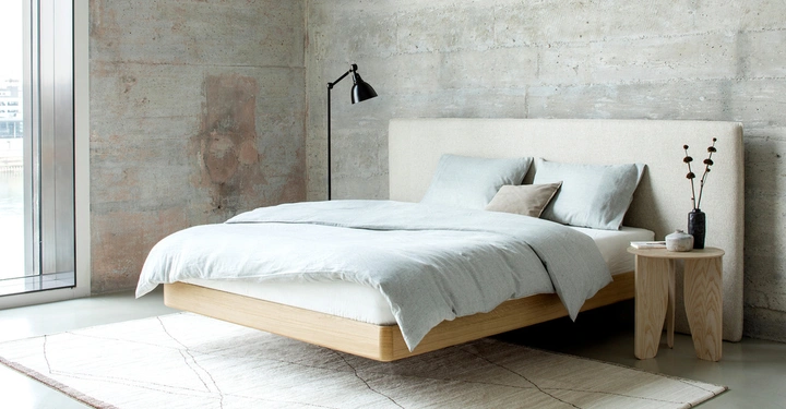 Bett mit grauer Bettwäsche und Kissen steht vor einer Betonwand in einem minimalistischen Schlafzimmer Ein kleiner Tisch mit Vase und Lampe steht daneben ein großes Fenster ist links zu sehen