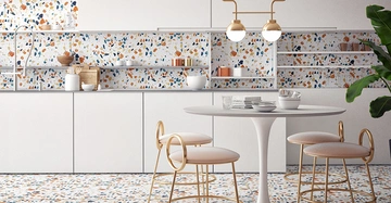 Ein runder Tisch mit goldenen Stühlen steht in einer modernen Küche mit weißen Schränken und bunten Terrazzo-Fliesen. Eine goldene Hängelampe leuchtet darüber, und eine Pflanze ist sichtbar.