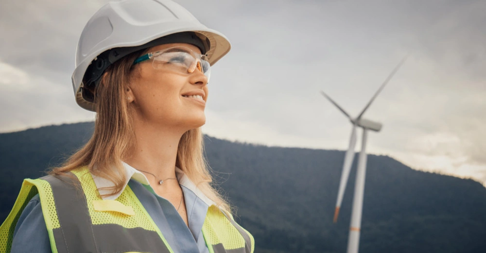 Junge blonde Frau in Warnweste und weißem Schutzhelm steht vor einer Windturbine und lächelt entschlossen in die Ferne.