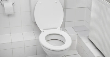 Weiße Toilettenschüssel mit offenem Sitz in einem gefliesten Badezimmer, daneben ein Toilettenpapierhalter und ein Heizkörper.