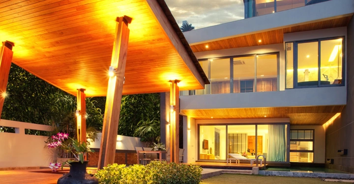 Ein modernes Haus mit großen Glasfenstern und abgestufter Architektur steht abends in einem beleuchteten Garten mit Holzüberdachung und Pflanzen an der Seite.