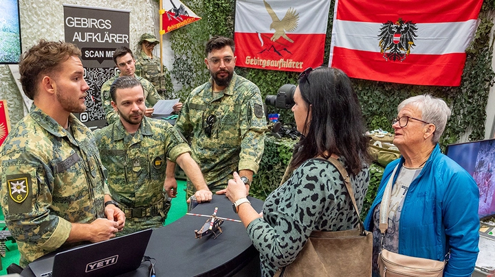 Galerie-Bild 4: Messestand der Gebirgsjäger mit vier Uniformierten und 2 Frauen, die sich mit ihnen unterhalten.