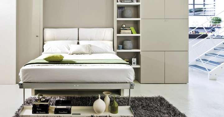 Bett mit weißen Kissen und Decke darauf liegendes grünes Kissen und Decke umgeben von weißen Regalen mit Dekoration und Büchern in einem modernen Zimmer Wohnzimmer im Hintergrund helle Treppe.