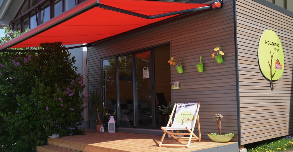 Holzgebäude mit roter Markise steht auf Terrasse neben Pflanzen Sitzstuhl und Schilder, ein Schild mit Text Holzhaus to go und Willkommen bei Anders! Holzhaus to go an der Wand unter Markise