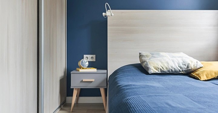 Schlafzimmer mit eisblauer Wand, davor ein weißes Boxspringbett mit blauer Tagesdecke und zwei Zierkissen in gelb und weiß. Neben dem Bett steht ein kleines Nachtkästchen.