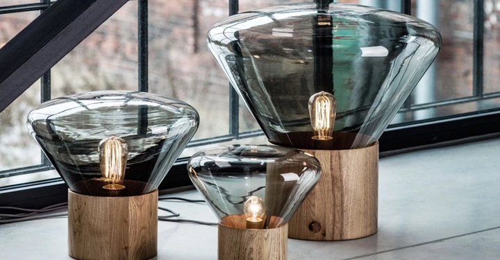 Galerie-Bild 6: Drei Tischlampen mit Holzsockeln und großen, gläsernen Schirmen stehen auf einer Betonfläche vor bodentiefen Fenstern in einem industriellen Loft. Die glühenden Glühbirnen sind sichtbar.