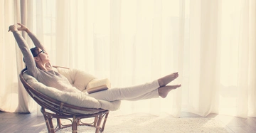 Person streckt sich entspannt in einem runden, gepolsterten Rattanstuhl mit einem offenen Buch auf den Knien in einem hellen, lichtdurchfluteten Raum mit weißen Vorhängen und einem weichen Teppich.