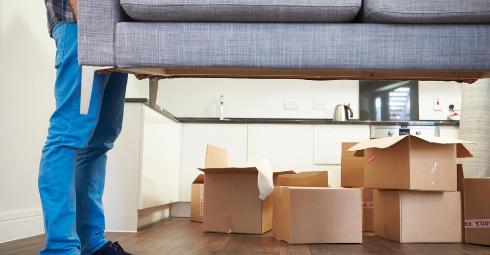 Jemand trägt ein graues Sofa in einer Wohnung, deren Küche im Hintergrund sichtbar ist. Umgeben von mehreren braunen Umzugskartons.