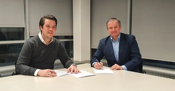 Zwei Männer sitzen in einem Büro an einem Tisch und unterzeichnen Dokumente. Im Hintergrund befinden sich große Fenster mit heruntergelassenen Jalousien.