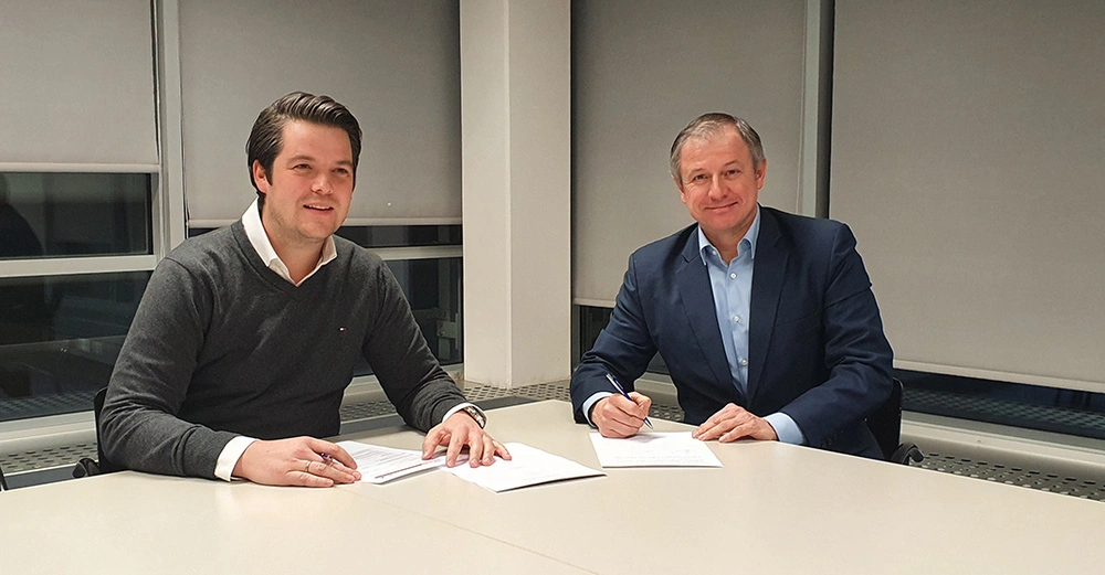 Zwei Männer sitzen in einem Büro an einem Tisch und unterzeichnen Dokumente. Im Hintergrund befinden sich große Fenster mit heruntergelassenen Jalousien.