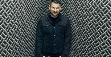 Mann trägt dunkle Jacke, lächelt, steht vor einem Hintergrund aus dicht aneinander liegenden, überlappenden Linien in einem geometrischen Muster.