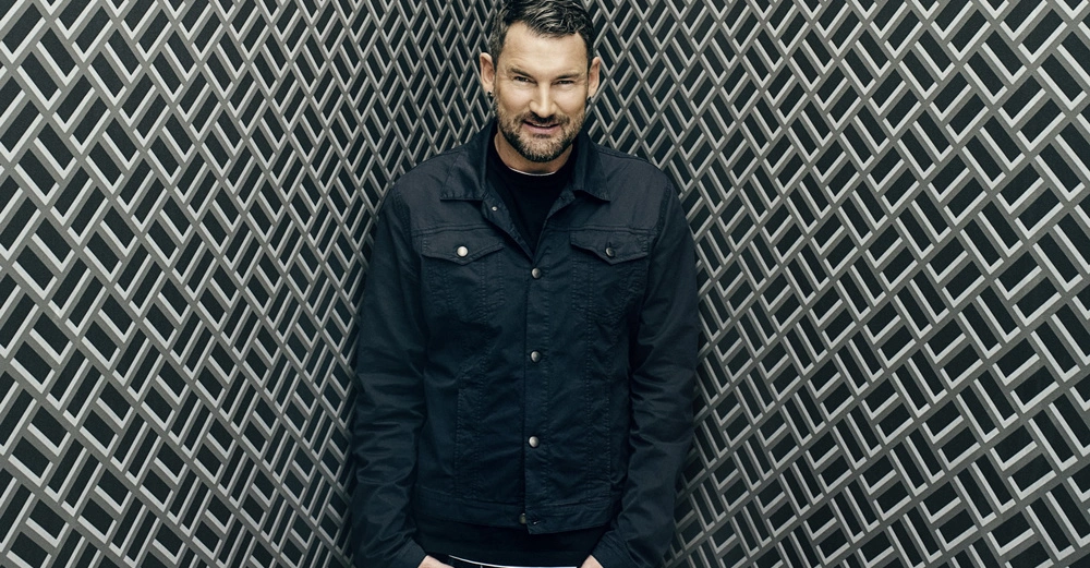 Mann trägt dunkle Jacke, lächelt, steht vor einem Hintergrund aus dicht aneinander liegenden, überlappenden Linien in einem geometrischen Muster.