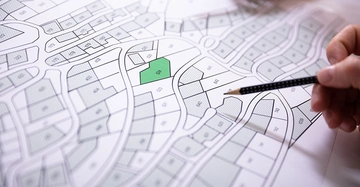 Eine Hand hält einen Bleistift und zeigt auf einen Stadtplan mit Grundstücksparzellen im Kontext einer urbanen Umgebung. Grundstück 524 ist grün markiert