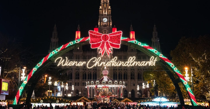 Beleuchteter Eingangsbogen zum Wiener Christkindlmarkt am Rathausplatz mit Karussell und Rathaus im Hintergrund.