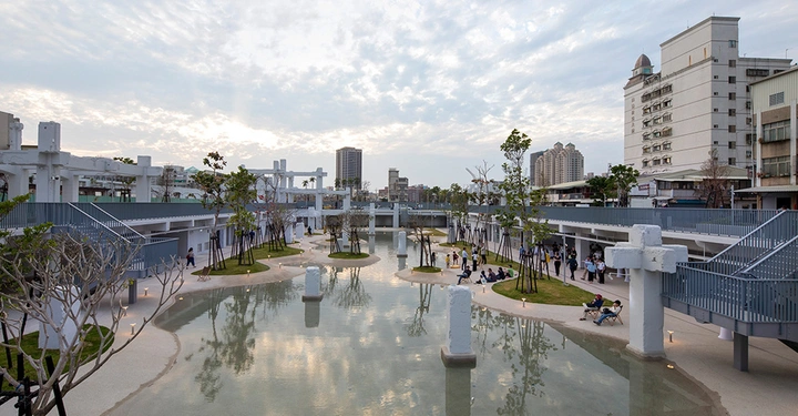Galerie-Bild 4: Menschen entspannen sich und spazieren um einen großen künstlichen Teich im Zentrum eines modernen Parks umgeben von Bäumen und Gehwegen in einer städtischen Umgebung bei bewölktem Himmel.