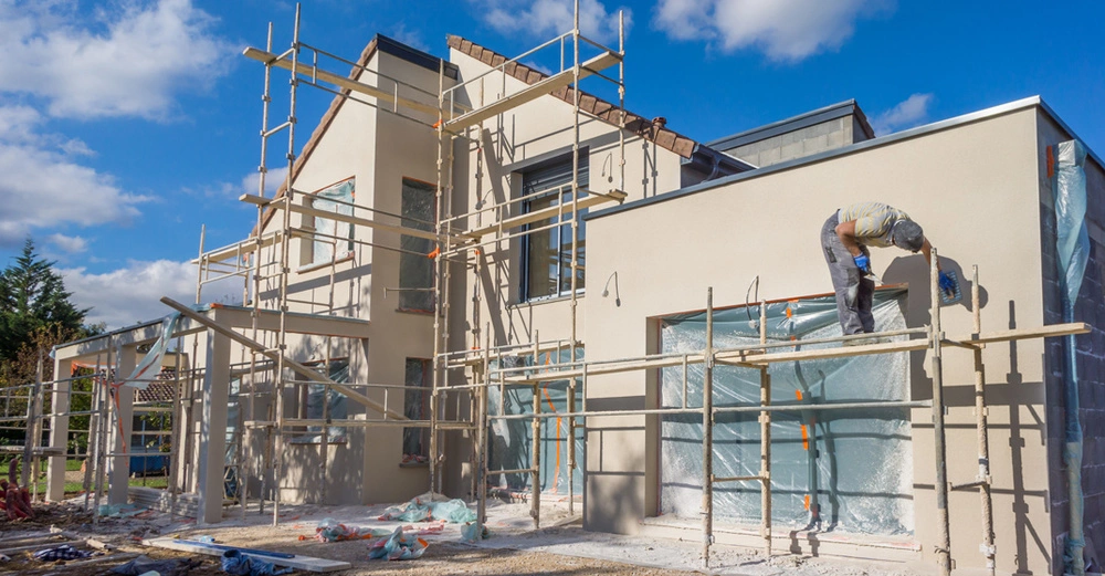modernes Einfamilienhaus mit Fassadengerüst. Auf dem Gerüst steht ein Maler und verputzt die Außenfassade in Sandfarbe. Blauer Himmel. 