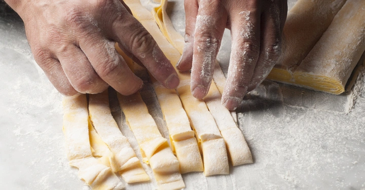 Hände schneiden frischen Pastateig in Streifen auf einer bemehlten Oberfläche in einer Küchenküchentheke.