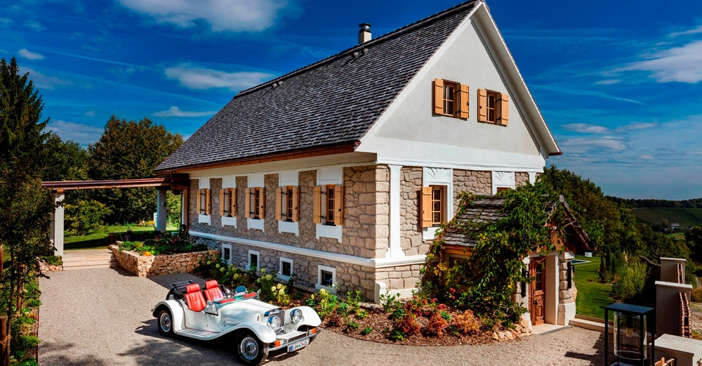 Ein Oldtimer mit offenem Verdeck und roter Innenausstattung parkt vor einem zweistöckigen Haus mit weißen Wänden und Holzbalken umgeben von einem gepflegten Garten in einer ländlichen Umgebung.