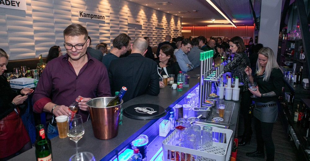 Der Mann im roten Hemd gießt ein Getränk ein an einer vollen Bar in einem belebten Raum beleuchtet mit neonfarbenen Lichtern. Im Hintergrund unterhalten sich Menschen. Schilder mit den Namen ADEY Kampmann Limot und Richter sind an der Wand befestigt