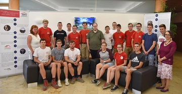 Gruppe Jugendlicher und Erwachsene posieren für ein Foto in einer modernen Lobby mit Sofas Fernseher und Info-Tafeln. Text auf Info-Tafel: Für die Geschichte von Hartl Haus 1980 1995 1997 1998 2001 2002 2005.