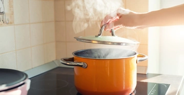 Hand hebt Deckel eines orangefarbenen Topfs auf Herdplatte an wobei Dampf aufsteigt umgeben von einer hellen Küchenumgebung mit Kacheln.