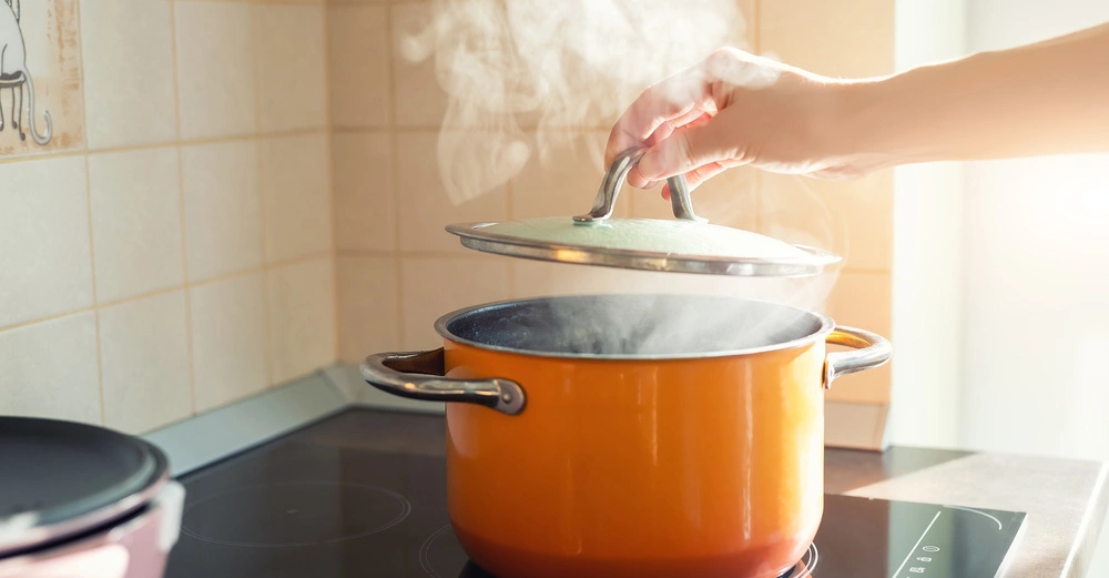 Hand hebt Deckel eines orangefarbenen Topfs auf Herdplatte an wobei Dampf aufsteigt umgeben von einer hellen Küchenumgebung mit Kacheln.