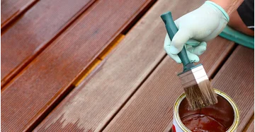 Eine Person mit Schutzhandschuh trägt mit einem Pinsel Holzlasur auf Terrassendielen auf. Neben dem Pinsel steht eine geöffnete Dose mit Holzschutzfarbe.