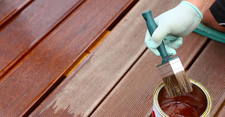 Eine Person mit Schutzhandschuh trägt mit einem Pinsel Holzlasur auf Terrassendielen auf. Neben dem Pinsel steht eine geöffnete Dose mit Holzschutzfarbe.