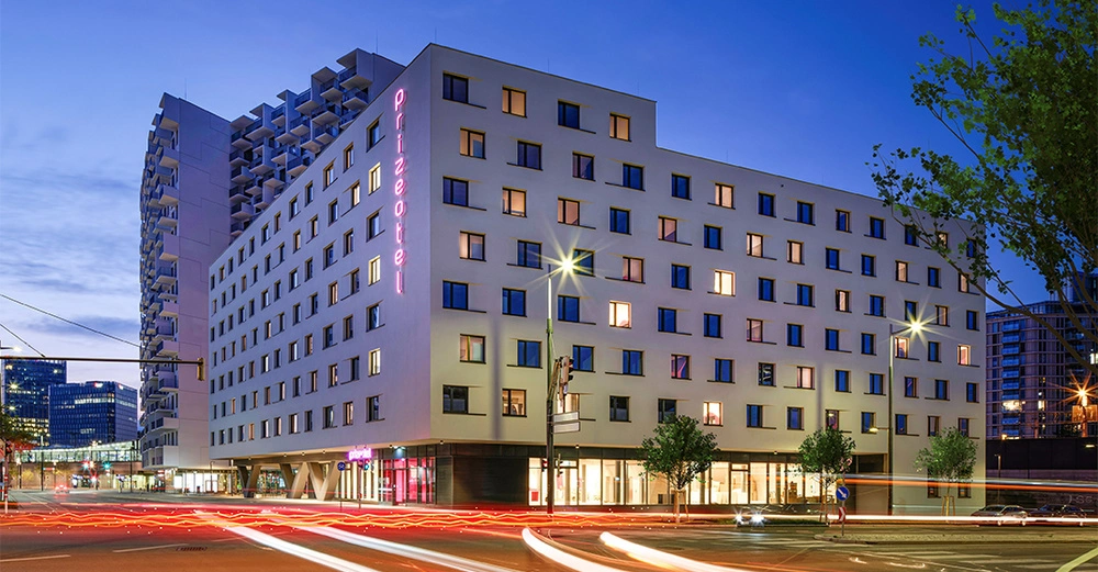 Rechteckiges Hotelgebäude leuchtet in der Dämmerung mit rosa Neonschriftzug Prizeotel Passanten und Autolichter bewegen sich in einer geschäftigen städtischen Umgebung mit Bäumen im Vordergrund.