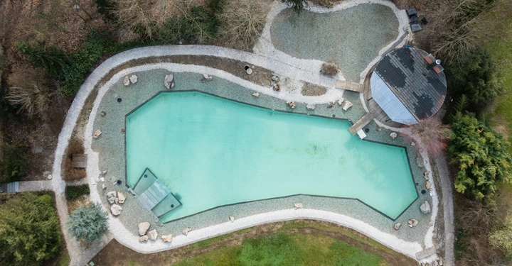 Ein grünliches Wasserbecken liegt inmitten einer kiesbedeckten Landschaft. Es gibt umliegende Bäume, Büsche und Pfade sowie ein rundes Gebäude mit einer Holzterrasse, die ins Wasser ragt.