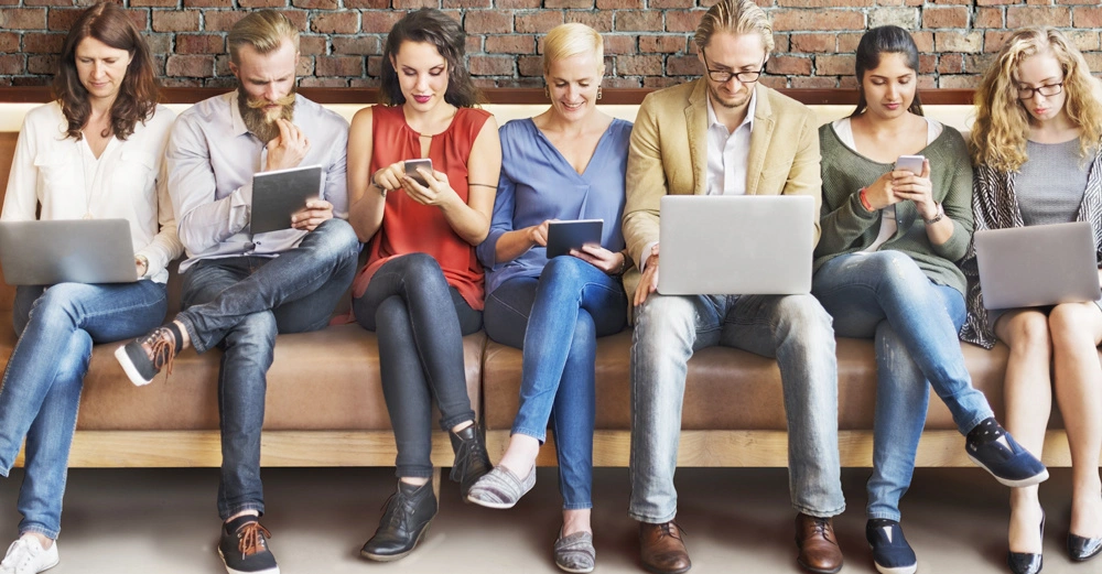 Personen verschiedener Altersgruppen sitzen auf einer Couch und nutzen verschiedene elektronische Geräte. Im Hintergrund eine Ziegelsteinmauer.