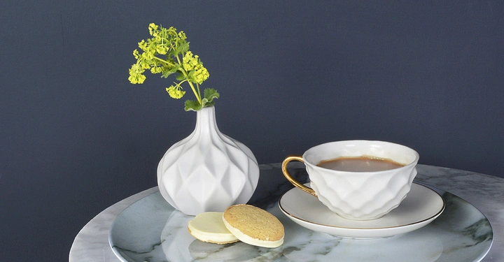 Galerie-Bild 1: Weiße Vase mit gelben Blumen steht neben einer Tasse Kaffee und zwei Keksen auf einem Marmor-Tablett. Der Hintergrund ist dunkelblau.