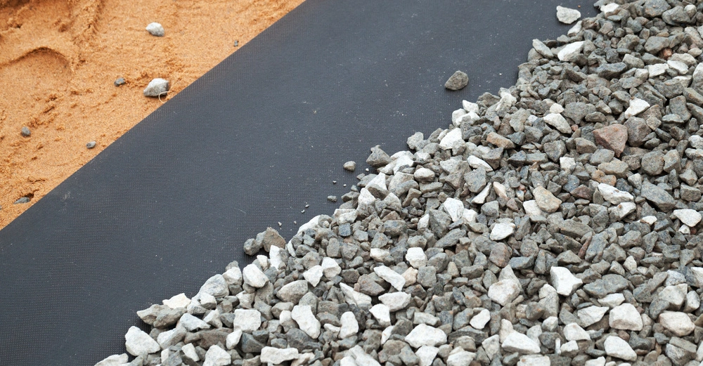 Kieselsteine liegen auf einem schwarzen Geotextilmaterial während auf der linken Seite Sand zu sehen ist.