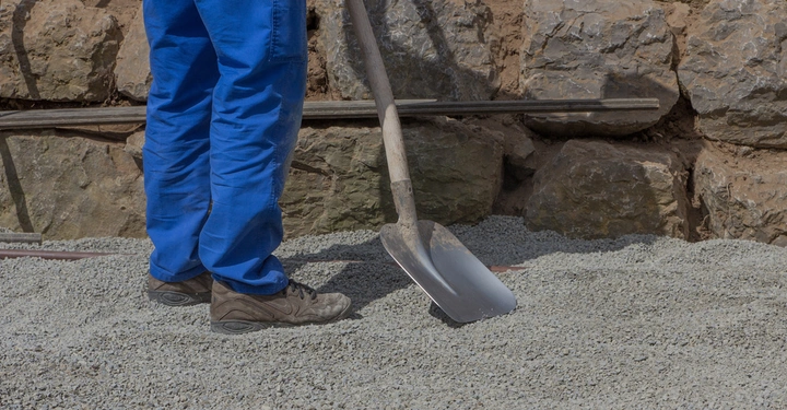 Eine Person in blauer Arbeitshose hält eine Schaufel auf einem Schotterhaufen, neben einer Steinmauer.