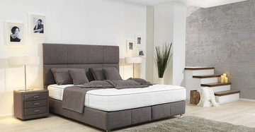 Grau bezogenes Bett steht mit mehreren Kissen und Decke in einem modern eingerichteten Schlafzimmer mit Wandbildern neben Nachttischen und Stehlampen; Treppe mit Dekoration in Ecke.