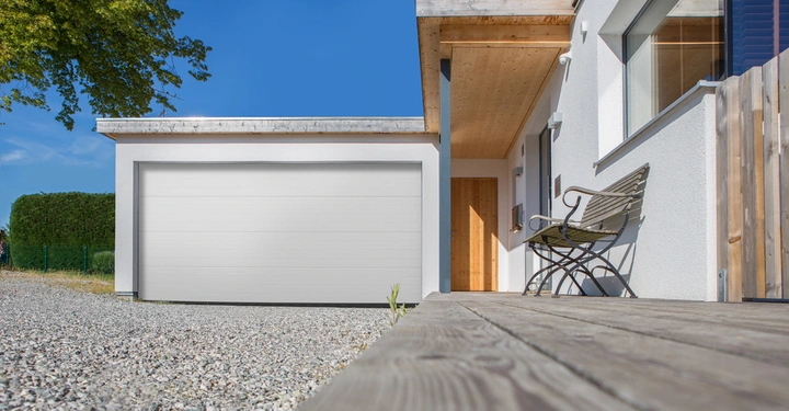Weiße Garage geschlossen stehend vor einer modernen Hausfassade mit Holztür und Fenster Bank daneben auf Holzterrasse davor Schotterweg mit grünem Baum und Hecke im Hintergrund blauer Himmel