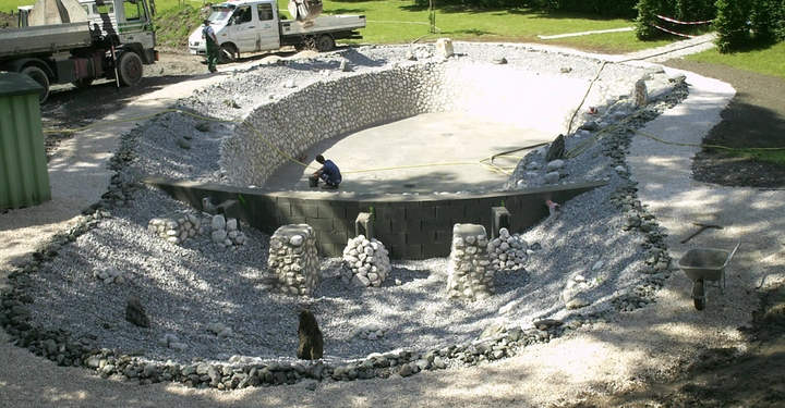 Person installiert Steinmauer stehend in großer kreisförmiger Baugrube umgeben von Kies während LKWs und Arbeiter auf Baustelle im Freien warten.