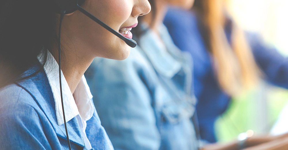 Eine Frau trägt ein Headset und spricht in einem Büro neben weiteren Personen in ähnlichen Positionen vor Computern oder Telefonen.