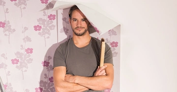 Ein Mann hält einen Pinsel und hat eine Tapetenbahn auf seinem Kopf in einem Raum mit blumengemusterter Tapete.