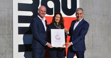 Drei Menschen halten ein gerahmtes Zertifikat und lächeln fröhlich vor einem Gebäude mit einem großen Baulogo Text: Baumit GmbH Beste Produktmarke Auszeichnung 2019.