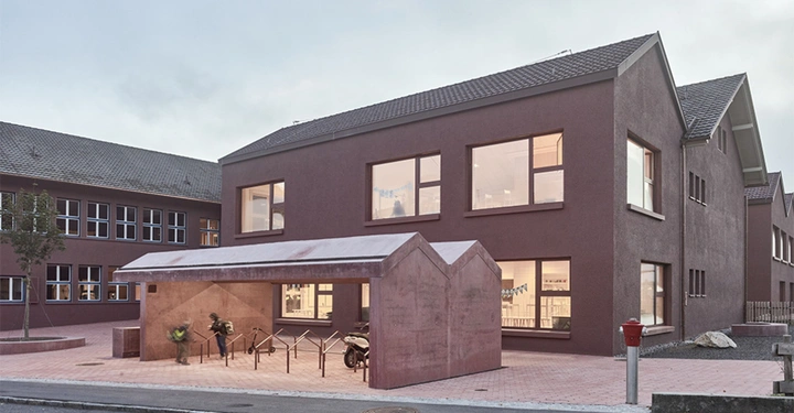 Rotes Gebäude mit großen Fenstern zwei Personen parken Fahrräder unter überdachtem Fahrradständer gepflasterter Steinbereich Bäume und weiteres Gebäude im Hintergrund.