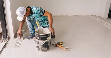 Handwerker verteilt Mörtel auf dem Boden einer renovierten Innenraumwohnung und kniet dabei neben einem Eimer mit Werkzeugen.