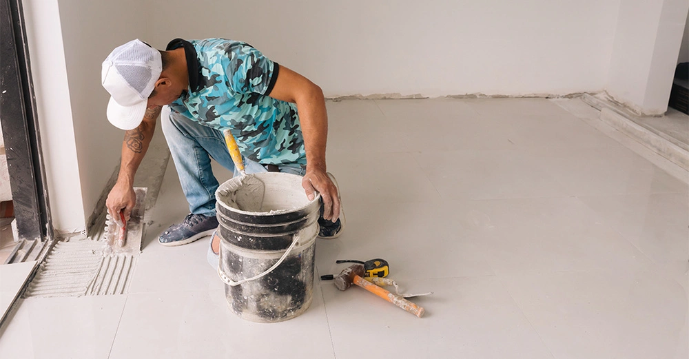 Handwerker verteilt Mörtel auf dem Boden einer renovierten Innenraumwohnung und kniet dabei neben einem Eimer mit Werkzeugen.