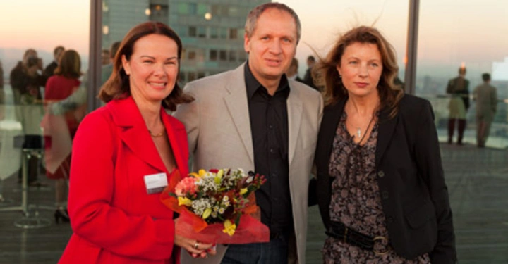 Galerie-Bild 33: Drei Personen stehen zusammen auf einer Terrasse bei Sonnenuntergang zwei Frauen und ein Mann die Frau links hält einen Blumenstrauß im Hintergrund sind Menschen und ein Hochhaus zu sehen.