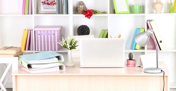 Ein Laptop steht auf einem Schreibtisch neben einer Schreibtischlampe im Vordergrund eines Regals mit Aktenordnern, Büchern und Dekorationen in einem Büro.