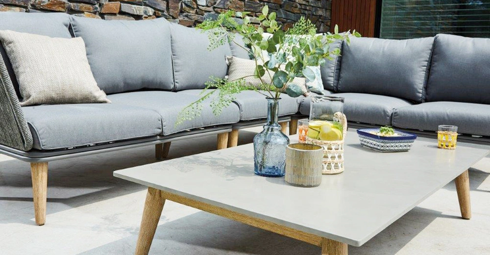 Graue Couch mit Kissen steht auf einer Terrasse. Ein Couchtisch mit Dekoration und Getränken im Vordergrund. Steinmauer im Hintergrund.