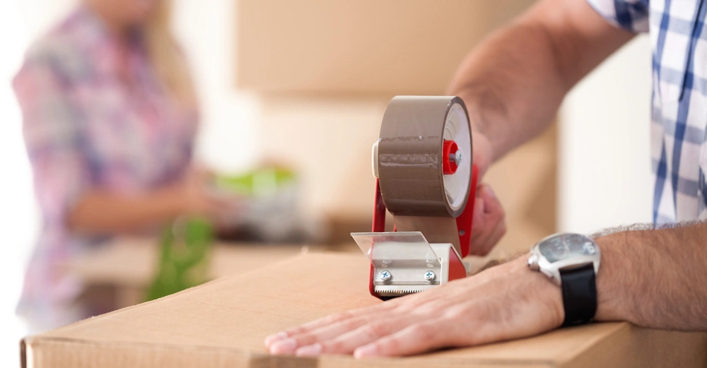 Person packt einen Karton mit Klebeband in einem Raum voller Kisten. Im Hintergrund hilft eine weitere Person.